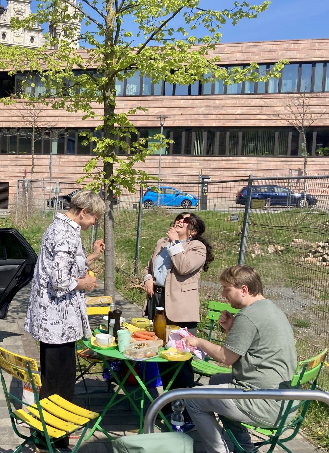Teamtreff im Freien: drei Menschen an einem grünen Bistrotisch mit Obst, Karotten und Saft, lachend in der Frühlingssonne